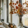 Moderne Herbstdeko für große Fenster mit Vase, Zweigen, Kürbissen und Kerzen auf heller Fensterbank.