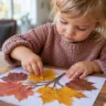 Basteln mit Kleinkindern im Herbst: Ein Kind gestaltet mit bunten Blättern ein herbstliches Bild am Holztisch.