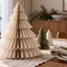Drei gefaltete Weihnachtsbäume aus cremefarbenem und grünem Papier schmücken einen festlich gedeckten Holztisch.