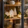 Moderne schwarze Glasvitrine mit weißen Weihnachtshäusern, Tannenbaum, Stern, goldenen Kugeln und LED-Lichtern.