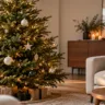 Moderner Weihnachtsbaum mit weißen, goldenen und dunkelgrünen Kugeln neben einem hellen Sofa im Wohnzimmer.