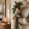 Weihnachtskranz aus beigen Leinenstreifen mit grünen Stoffbändern und Tannenzweigen an einer hellen Wand.