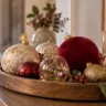 Rote, goldene und cremefarbene Weihnachtskugeln auf einem Holztablett mit Kerze und grünen Zweigen.