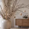 Große Keramikvase mit Birkenzweigen, Weihnachtsanhängern und warmweißer Lichterkette im Wohnzimmer.