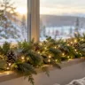 Breite Fensterbank mit Tannenzweigen, warmweißer Lichterkette und Zapfen vor einer verschneiten Landschaft.