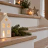 Moderne Weihnachtsdeko für Treppenstufen mit Keramikhaus, Tannengrün und warmem Licht.